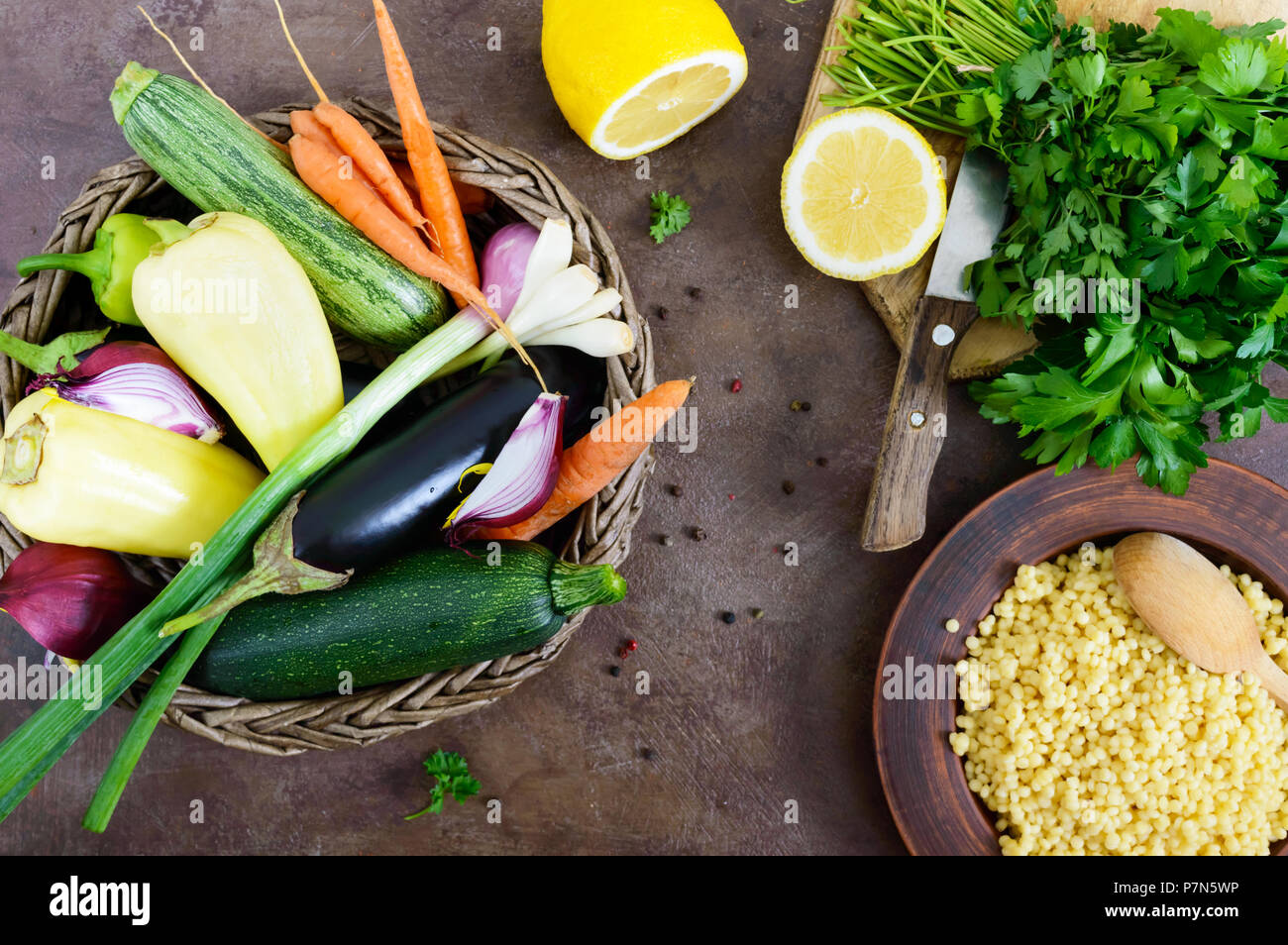 Delicious useful porridge cous cous in a clay plate and raw fresh vegetables in a basket, greens, lemon. Ingredients for cooking Tabbouleh. Vegetarian Stock Photo