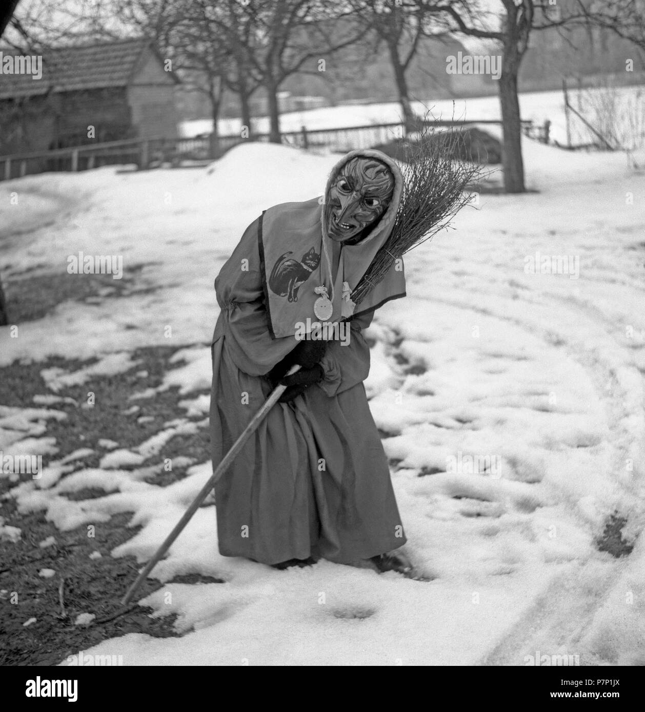 Witch with broom in hand stands in a curvilinear position in the snow, carnival, around 1950, near Freiburg, Germany Stock Photo