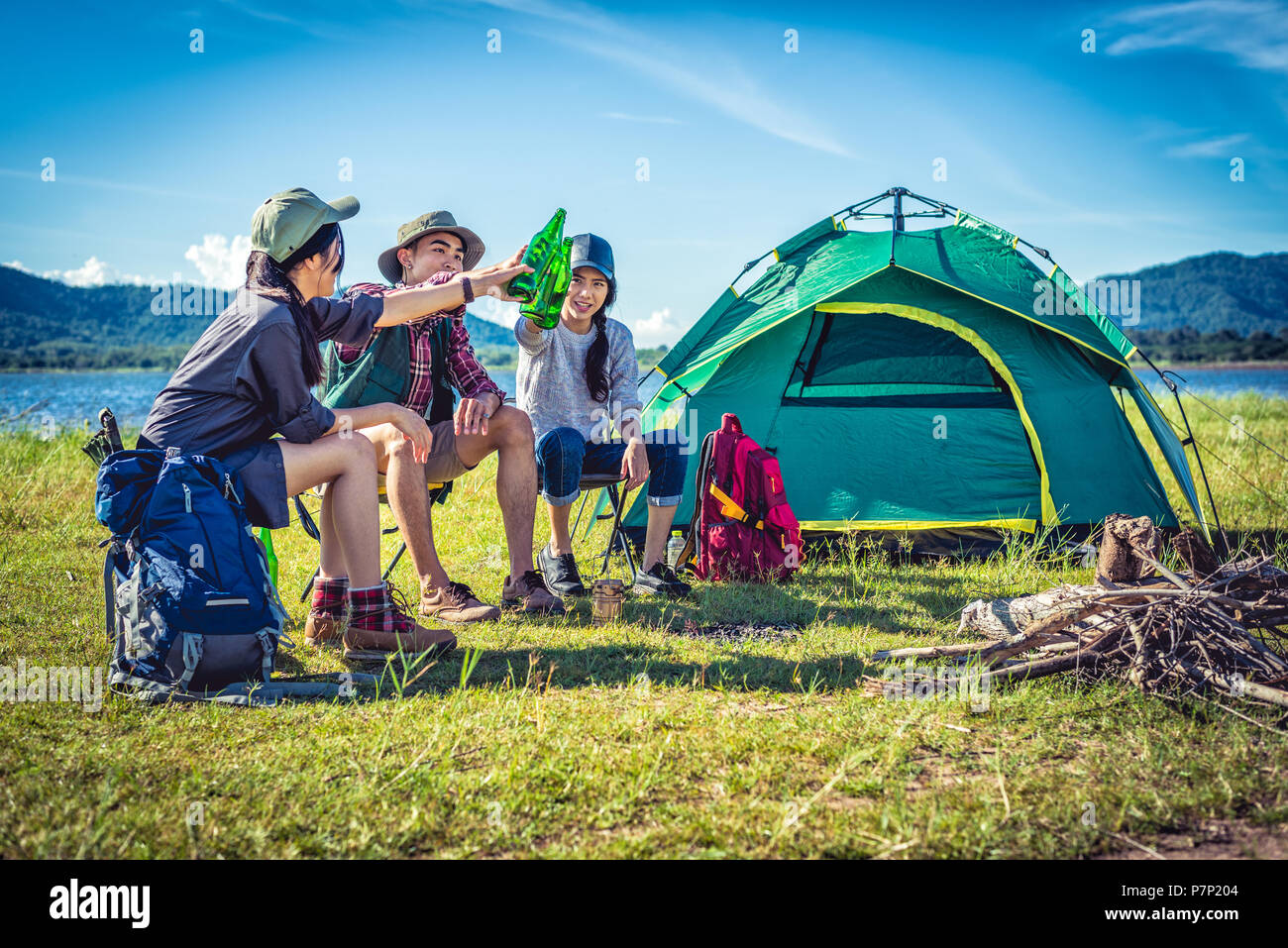 Group of young Asian friends enjoy picnic and party at lake with camping backpack and chair. Young people toasting and cheering bottles of beer. Peopl Stock Photo