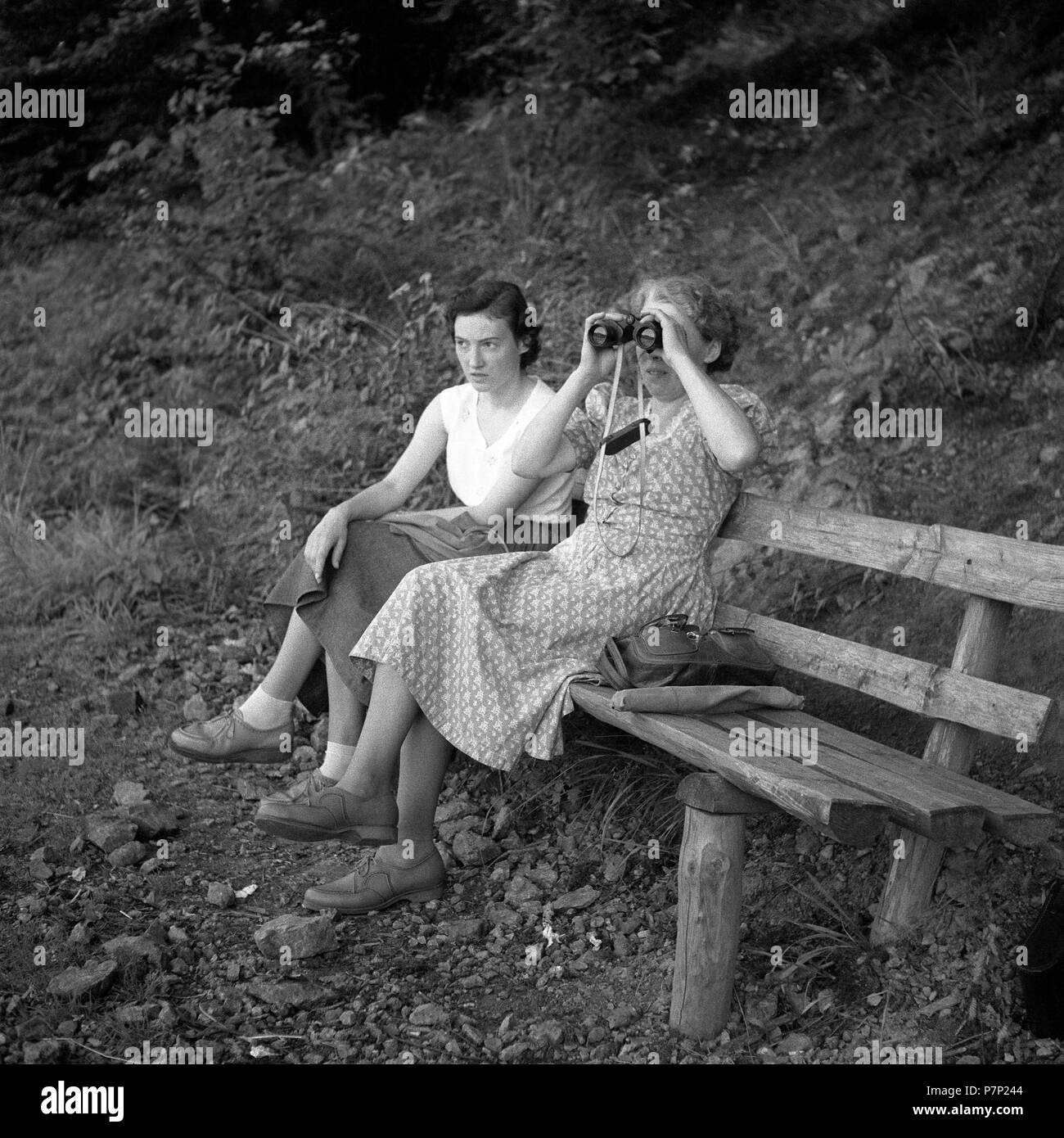 Two women are sitting on a bench and one of them is looking through binoculars, ca. 1945 to 1955, near Freiburg, Germany Stock Photo
