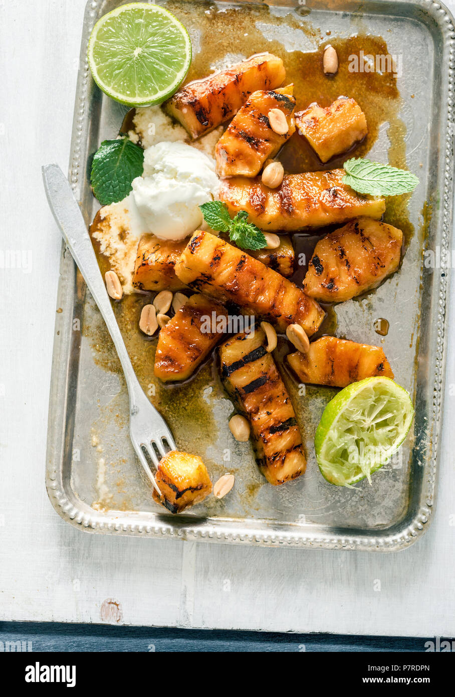 Tray of caramelized grilled pineapple with ice cream, mint, and nuts on a light wooden surface, top view Stock Photo