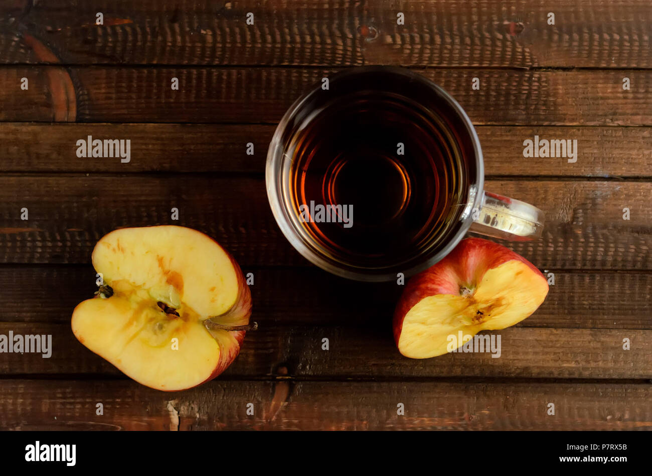 The glass of apple cider vinegar and two red halves of the apple on wooden background. Two red halves of the apple darkens on the cut. Flat lay view. Stock Photo