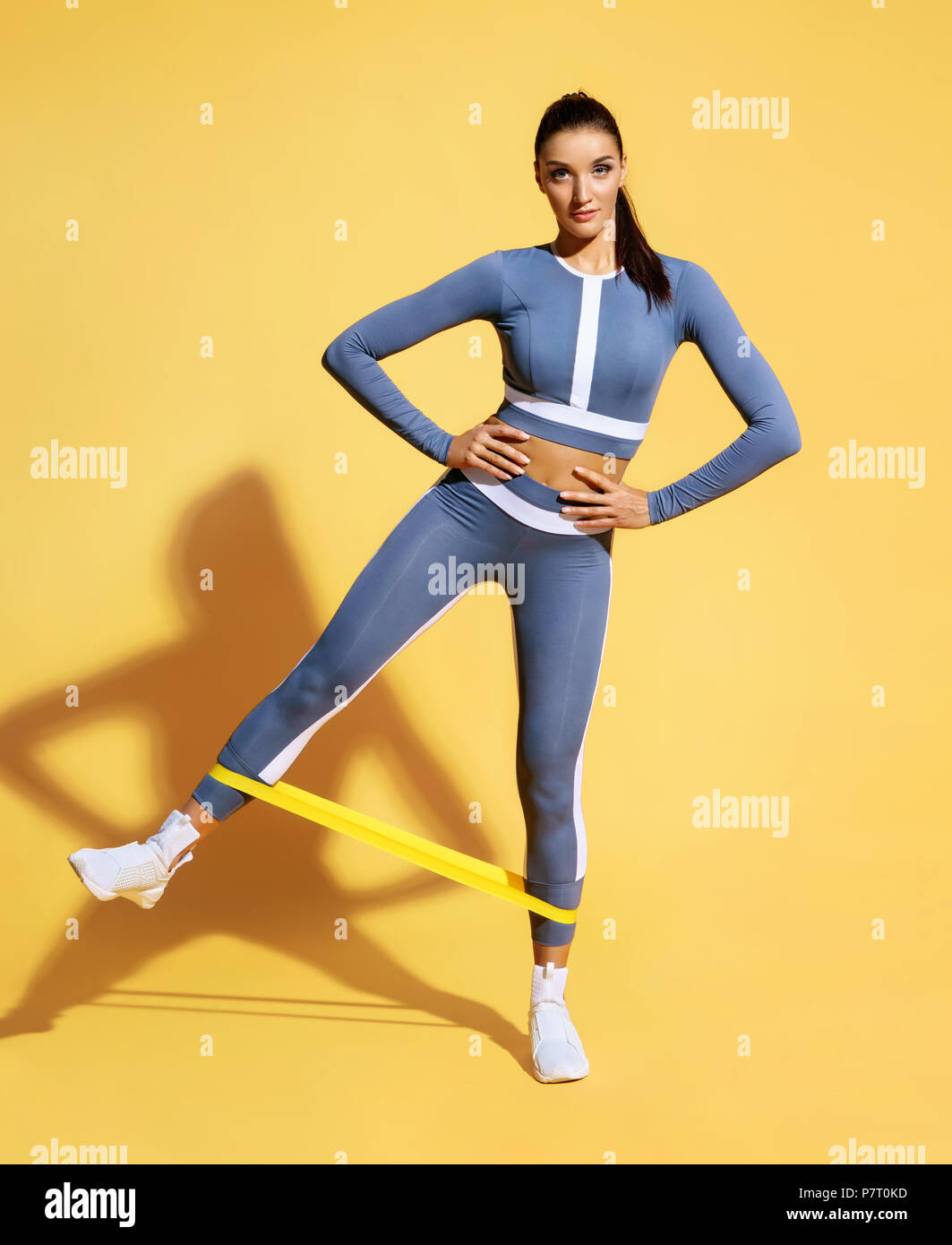 Woman with good physique doing stretching work out with elastic bands. Photo of latin woman in fashionable sportswear on yellow background. Strength a Stock Photo