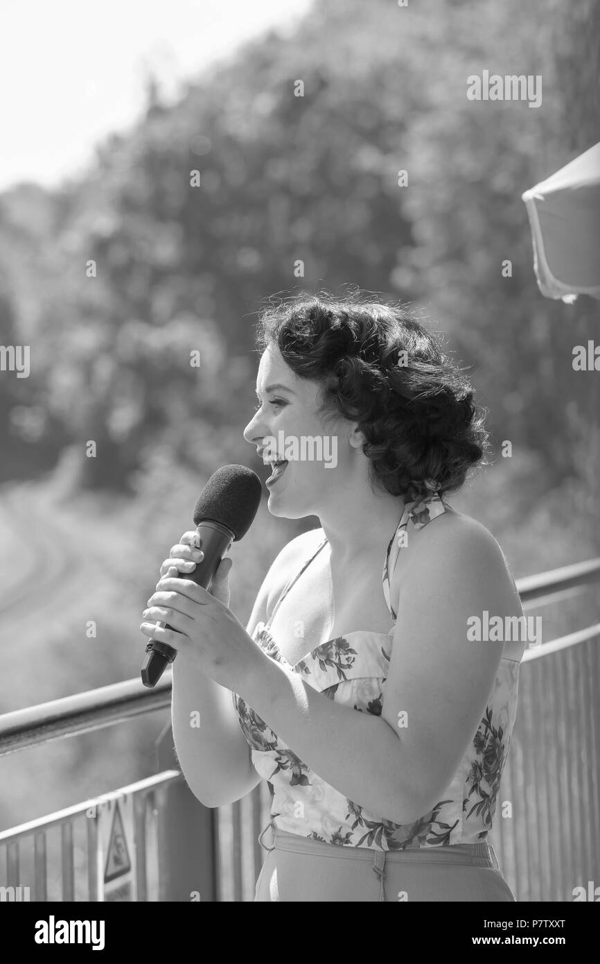 Kidderminster, UK. 7th July, 2018. A journey back in time continues at the Severn Valley Railway as we turn the clock back to the 1940s. Visitors and staff pull out all the stops to ensure a realistic second world war  Britain is experienced on this heritage railway line. Vintage singer, Hattie Bee entertains the crowds, pictured here singing into microphone, mouth open, in summer sunshine, on balcony overlooking railway line. Credit: Lee Hudson/Alamy Live News Stock Photo