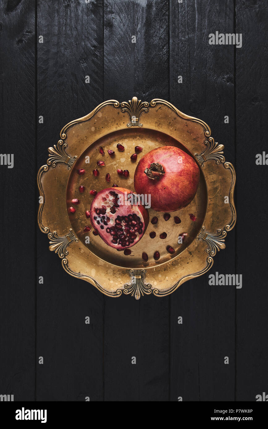 Flat lay of ripe pomegranate fruit surrounded by seeds on an old golden serving plate, black wooden vintage background. Top view with copy space. Stock Photo