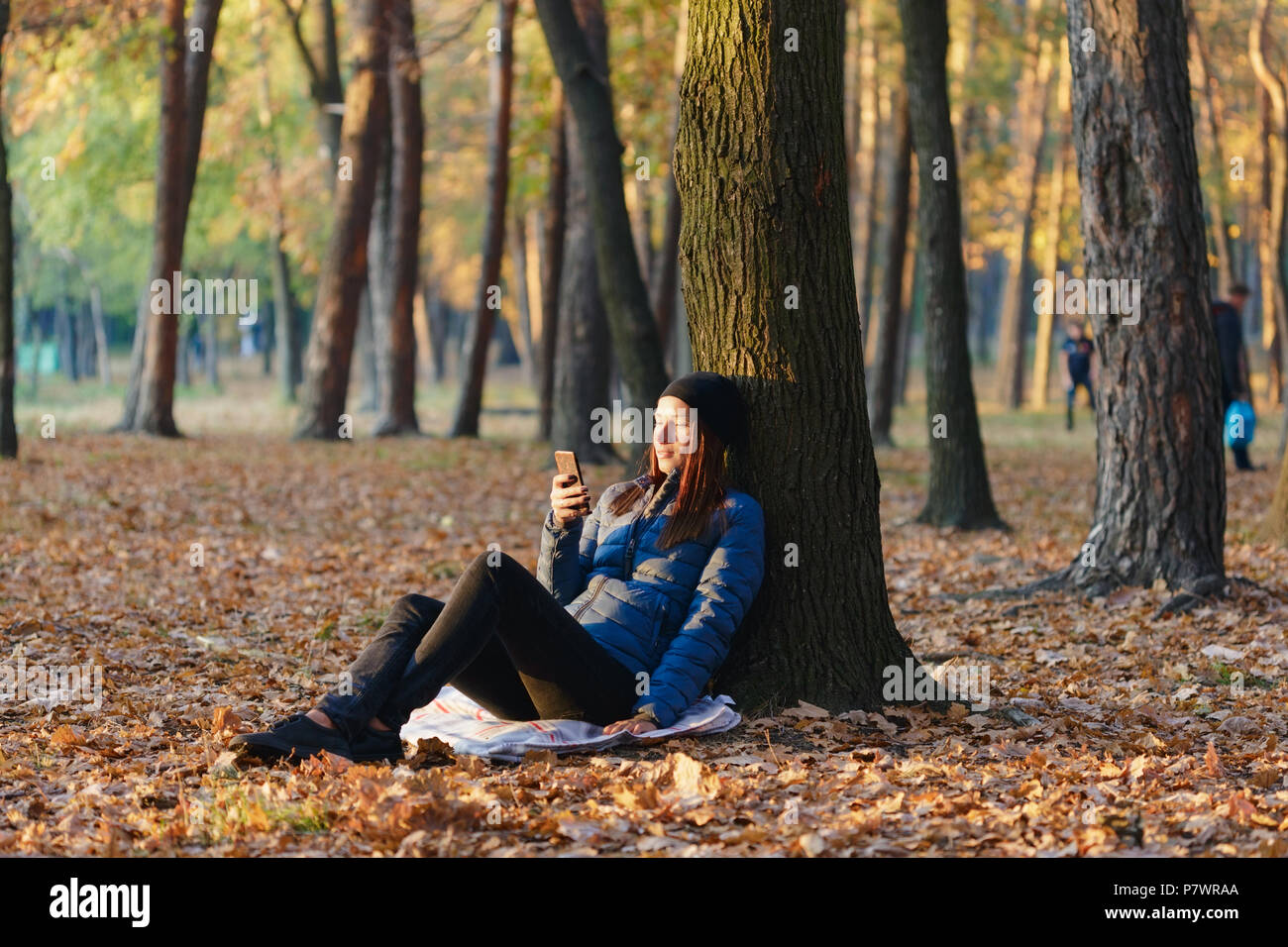 Autumn woman under a tree in autumn park communicates via the Internet using a smartphone Stock Photo