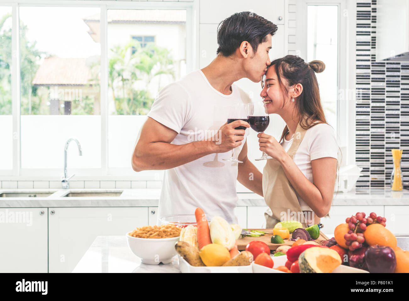 Asian lovers or couples kissing forehead and drinking wine in kitchen room at home. Love and happiness concept Sweet honeymoon and Valentine day theme Stock Photo
