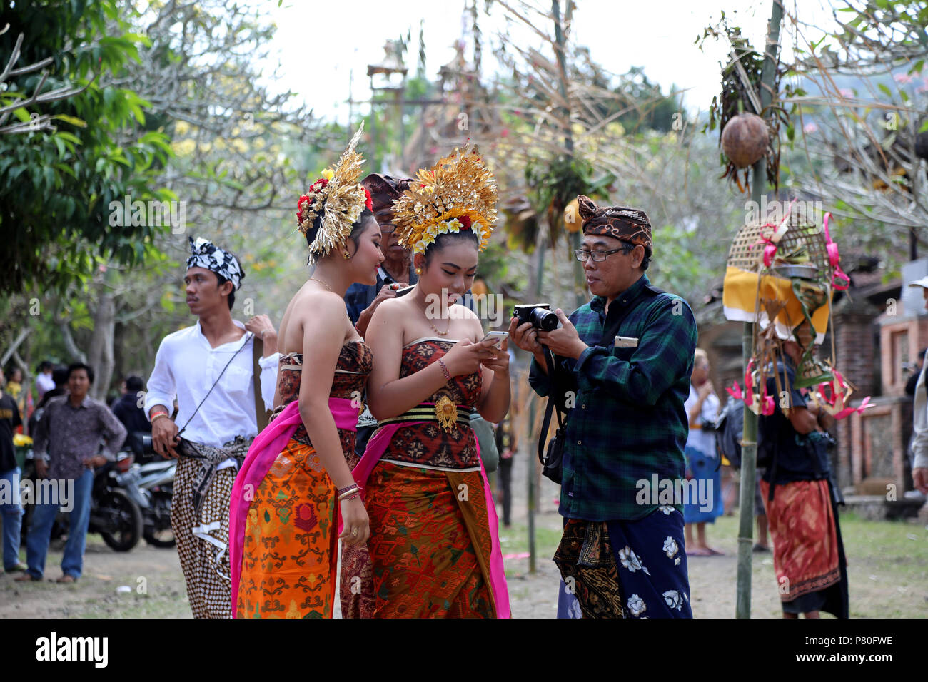 Tenganan, Indonesia – June 30 2018: Two local girls with ornate gold head-dresses gather around a mobile phone in the village of Tenganan, Bali during Stock Photo