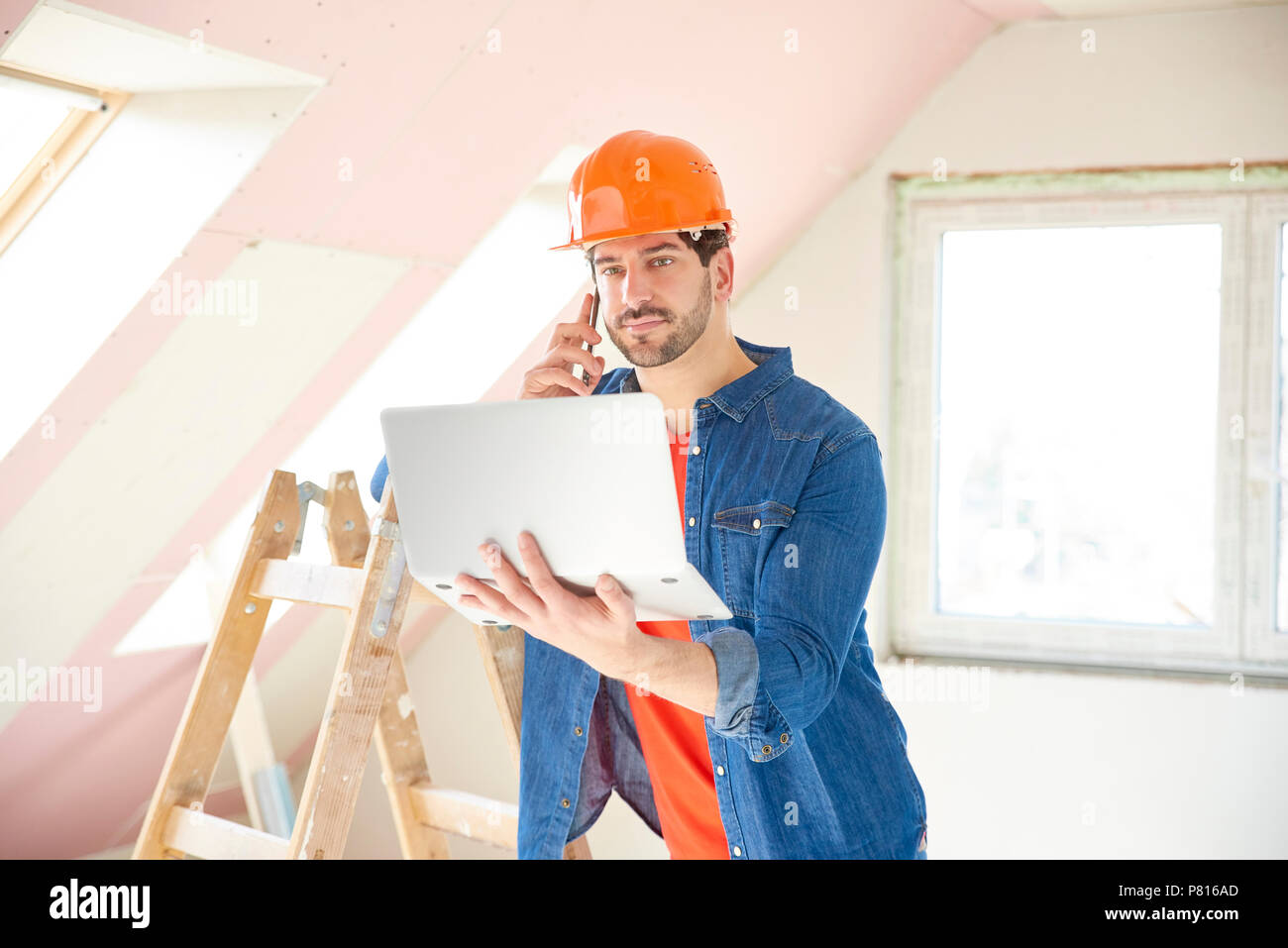 Young repairman using laptop and mobile phone while standing at renovation area. Stock Photo