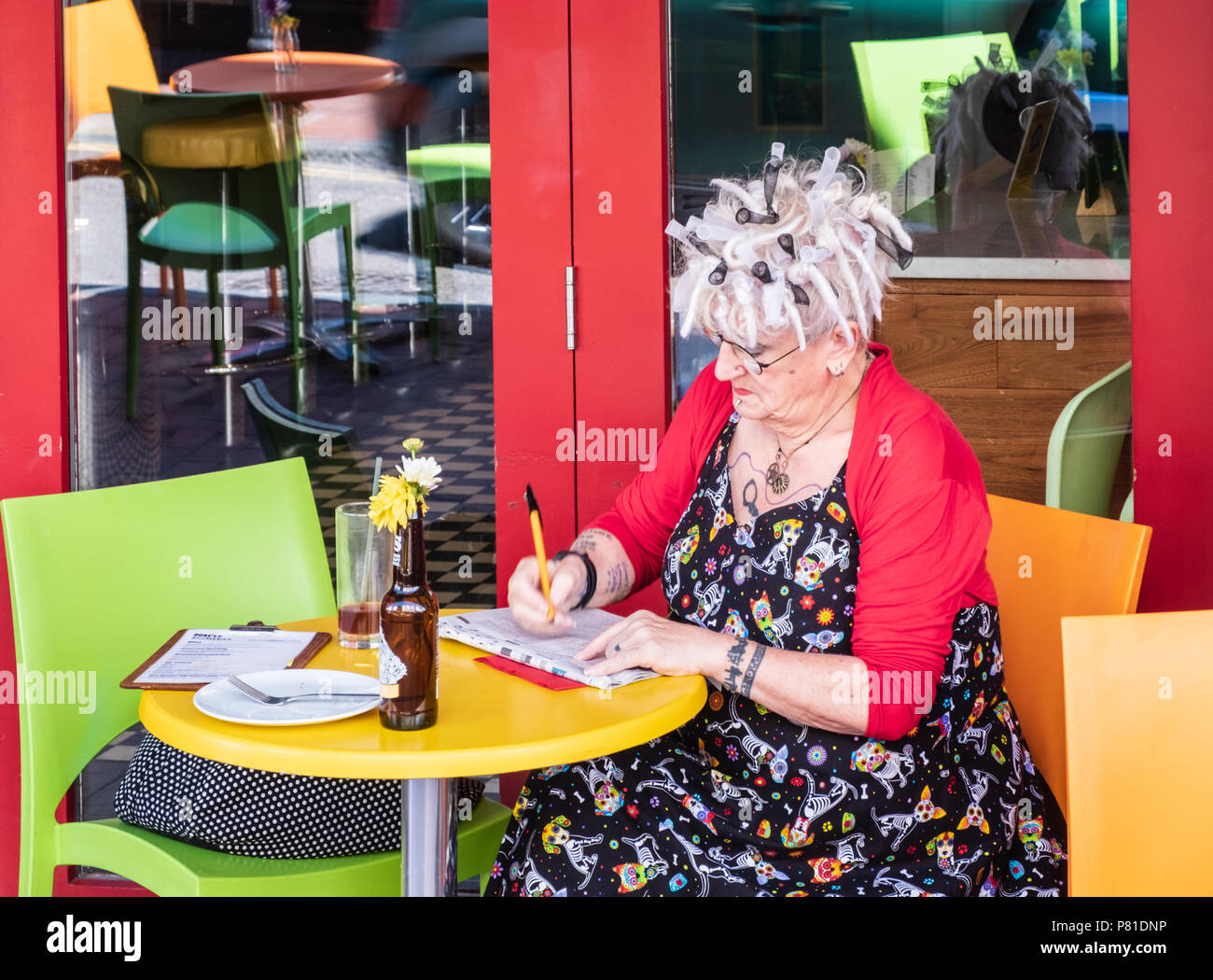 Free spirited older, stylish woman with tattoos and dressed in bright primary colours doing a crossword outside a cafe in Brighton, East Sussex Stock Photo