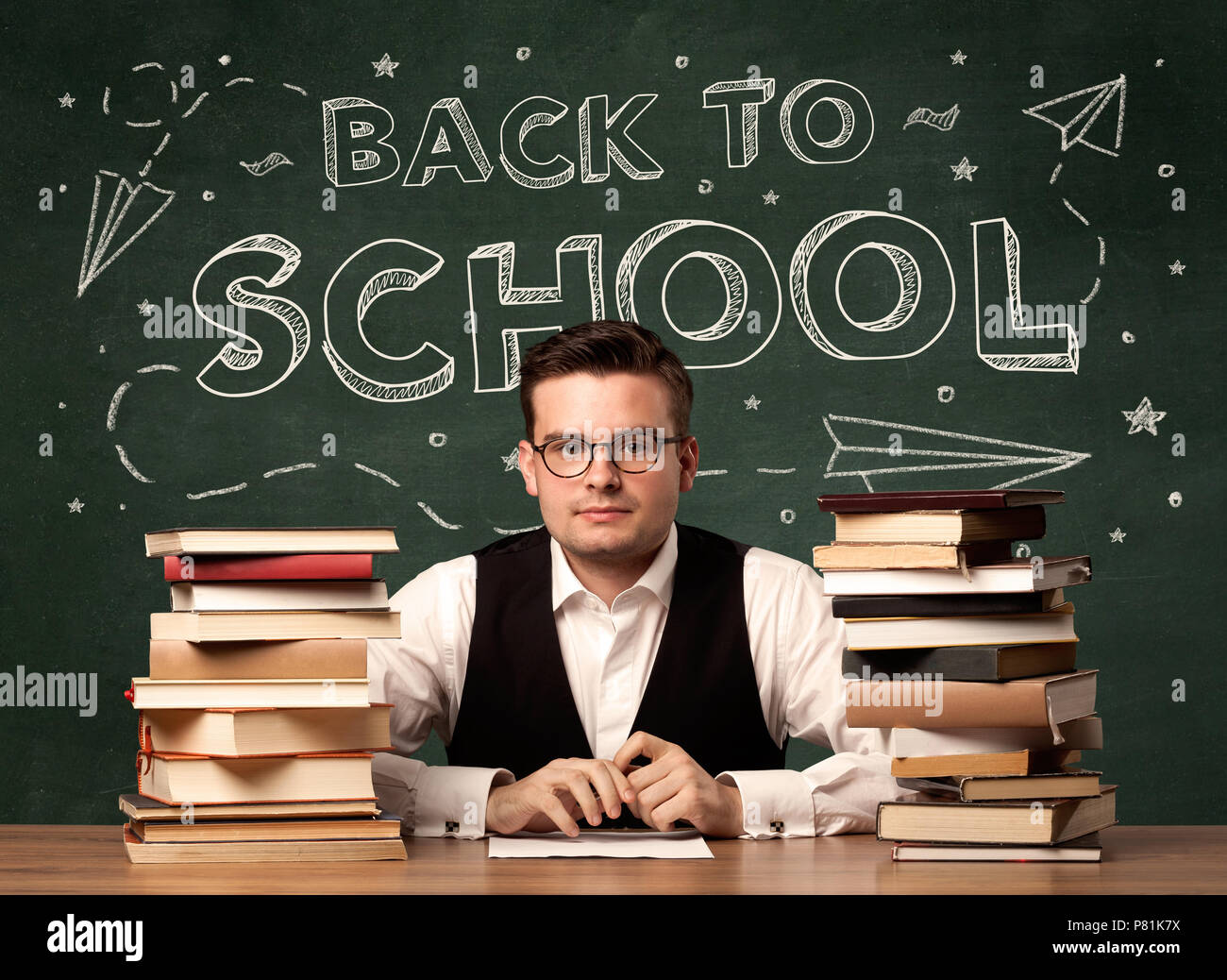 A young teacher in glasses sitting at classroom desk with pile of books ...
