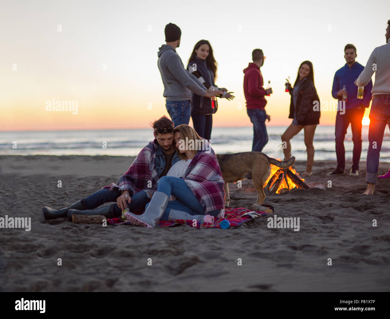 Young Couple Sitting with friends Around Campfire on The Beach At sunset drinking beer Stock Photo