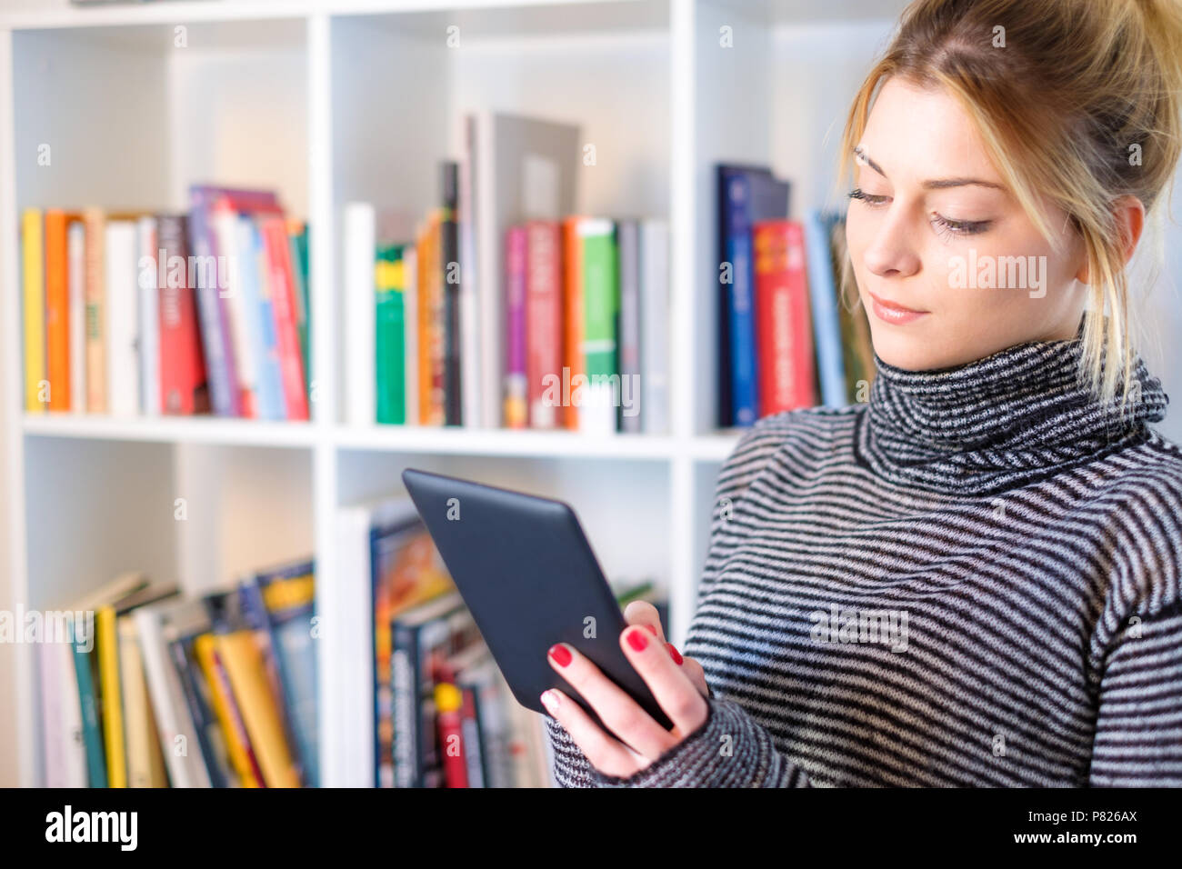 Young girl reading ebook reader next to colorful bookshelf Stock Photo