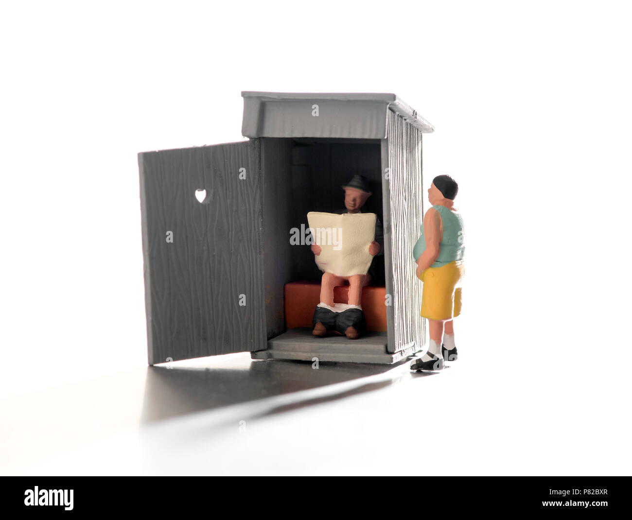 Fun scene of miniature people using an outdoor toilet with one person sitting reading inside the hut with the door open as a second waits impatiently  Stock Photo