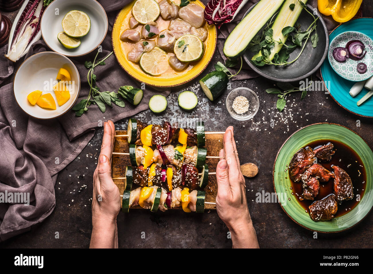 Female hands holding various homemade meat vegetables skewers for grill or bbq on rustic background with ingredients , plates and kitchen tools, top v Stock Photo