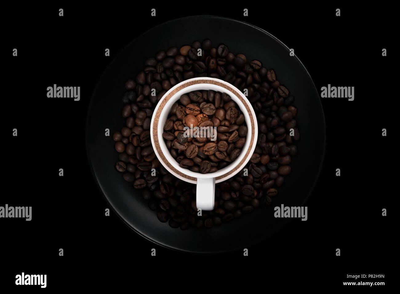 White cup full of coffee beans on black plate filled with roasted coffee beans. With negative space on black background. Stock Photo