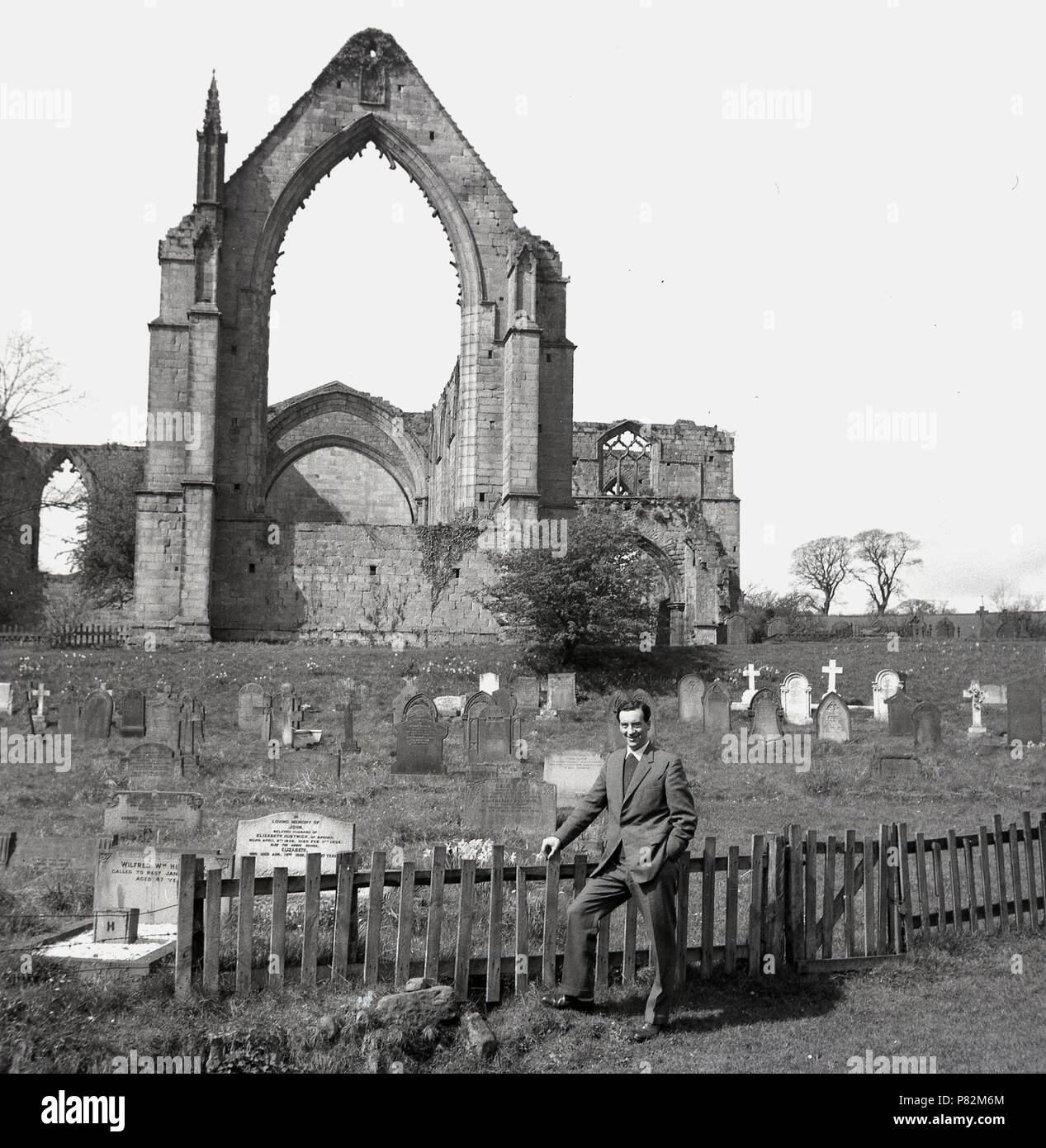 1959, historical, a man standing by a small wooden fence by the cementry which lies infront of the ruins of Bolton Abbey in Wharfedale, Yorkshire, England, UK.  The Augustinian Prioy ruins ly beside the river Wharfe. Stock Photo