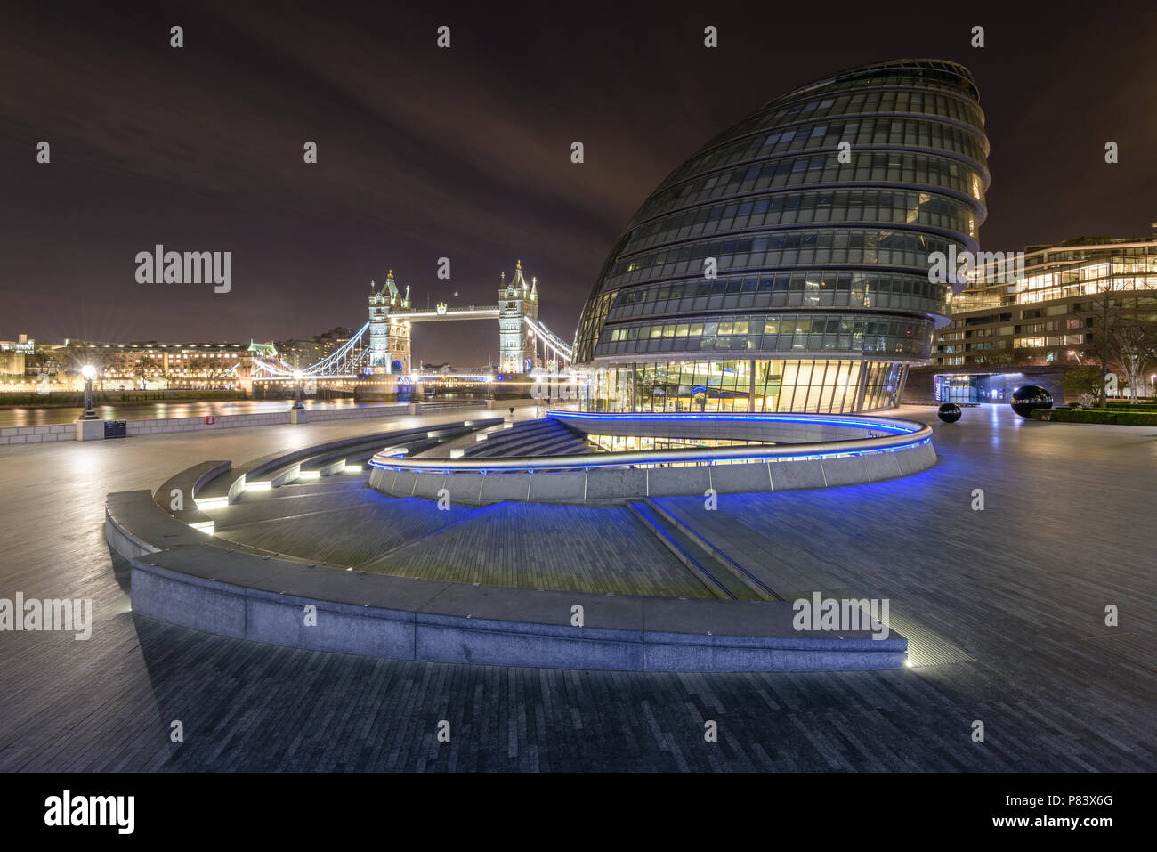 Meeting place at The Scoop amphitheatre at More London with City Hall and Tower Bridge lit up in blue and orange lights by the River Thames at night Stock Photo