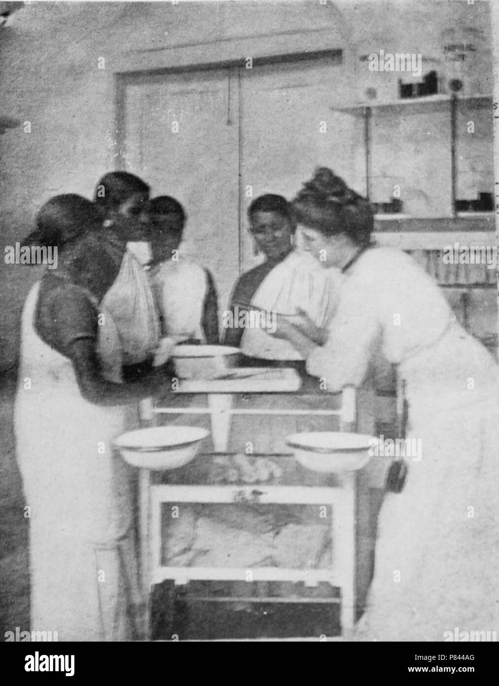 Black and white print depicting Miss ME McDonnell, wearing a Victorian dress and leaning over a table while speaking with a group of local nurses wearing saris, likely photographed at the CSI Nursing College, in Neyyoor, Tamil Nadu, India, 1912. Courtesy Internet Archive. () Stock Photo