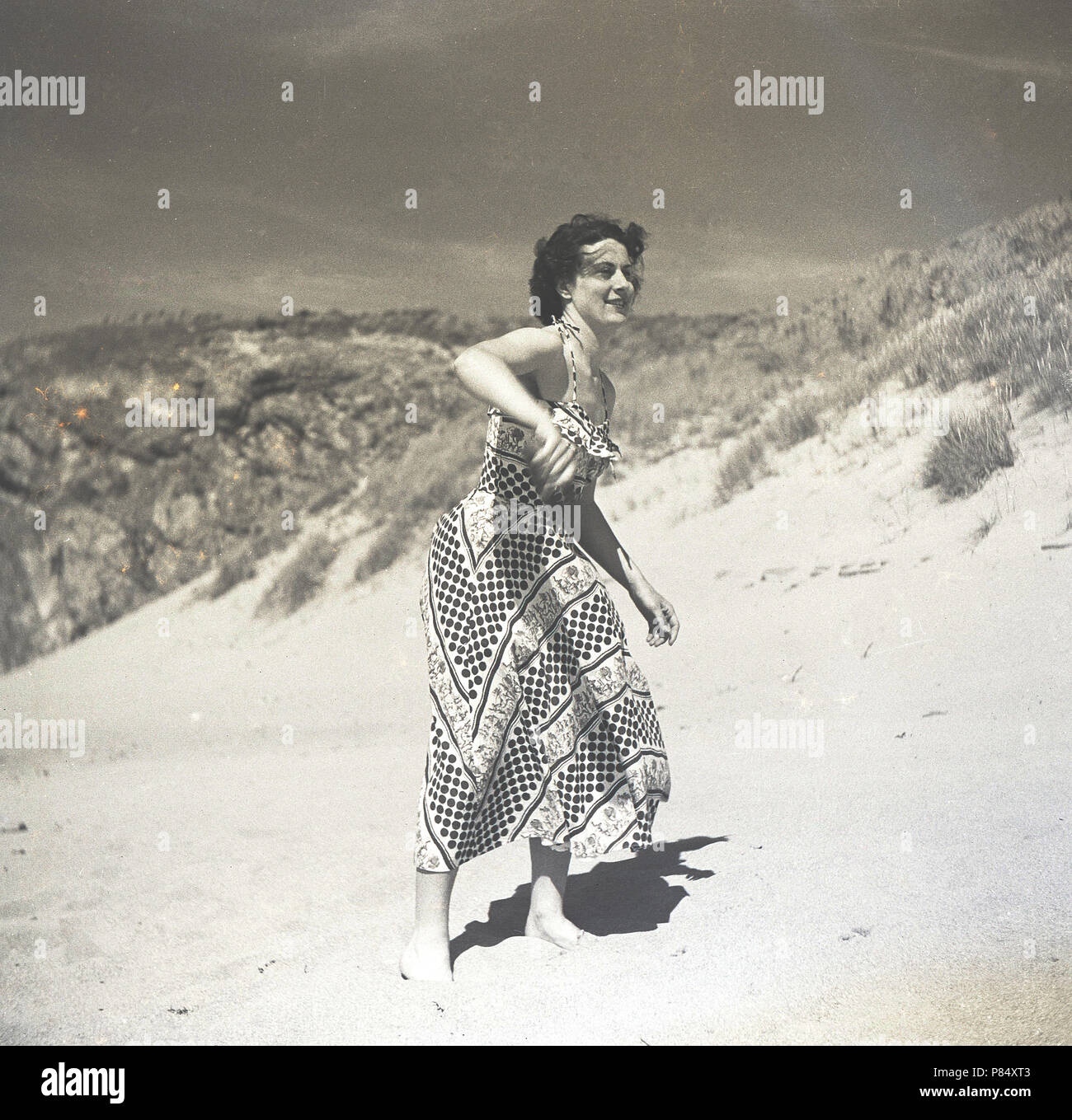 1950s, summertime and a young lady in a long patterned dress playing frisbee on a sandy beach, England, UK. Stock Photo