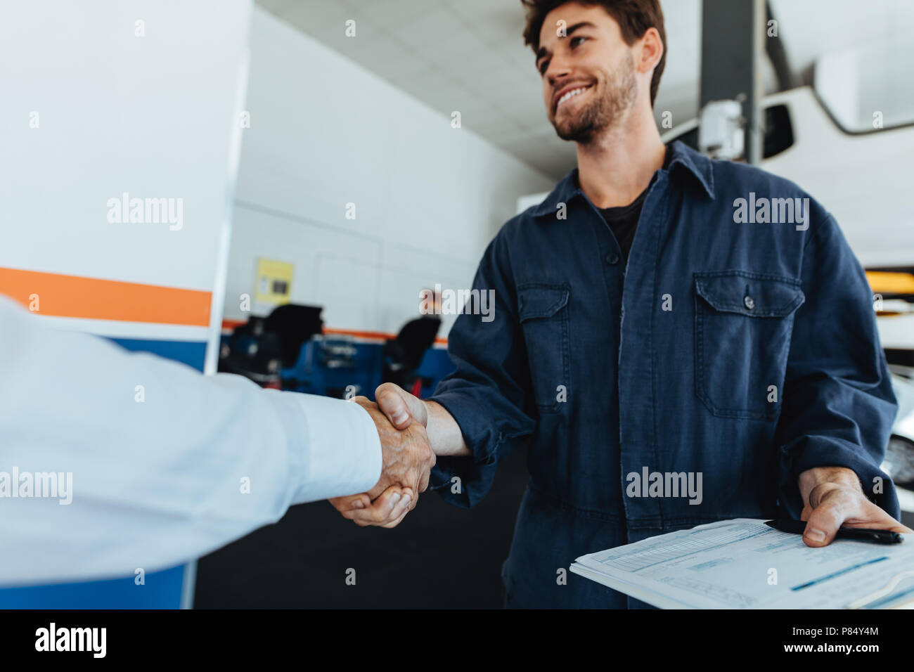 Young auto mechanic shaking hands with satisfied customer in garage. Automobile service center worker shaking hands with client after car servicing. Stock Photo
