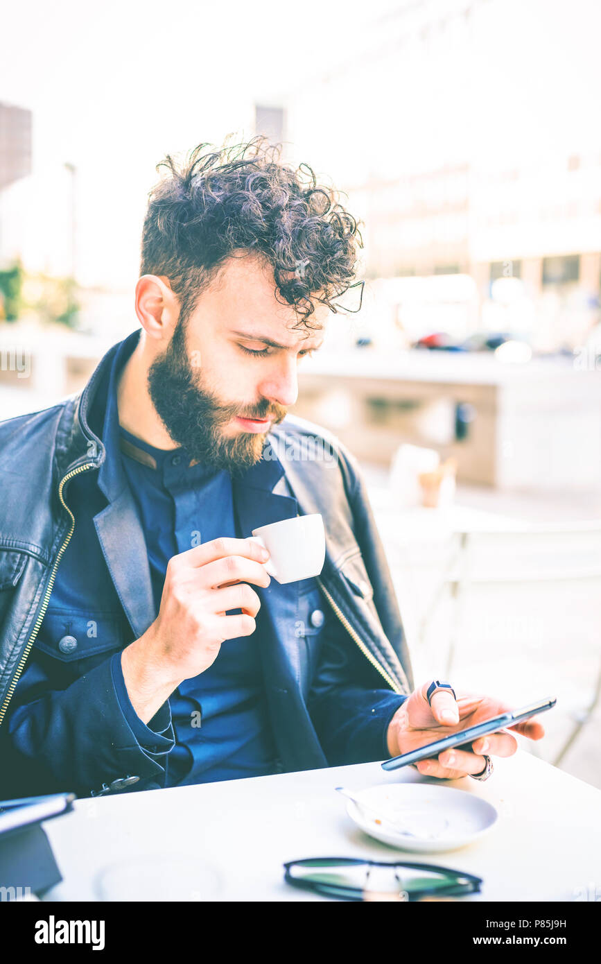 young handsome black hair caucasian modern businessman sitting in a bar, drinking coffee, using a smartphone - business, working concept Stock Photo