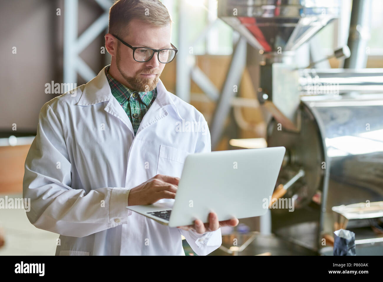 Waist up portrait of modern young man wearing lab coat using laptop while working in coffee production manufactory, copy space Stock Photo