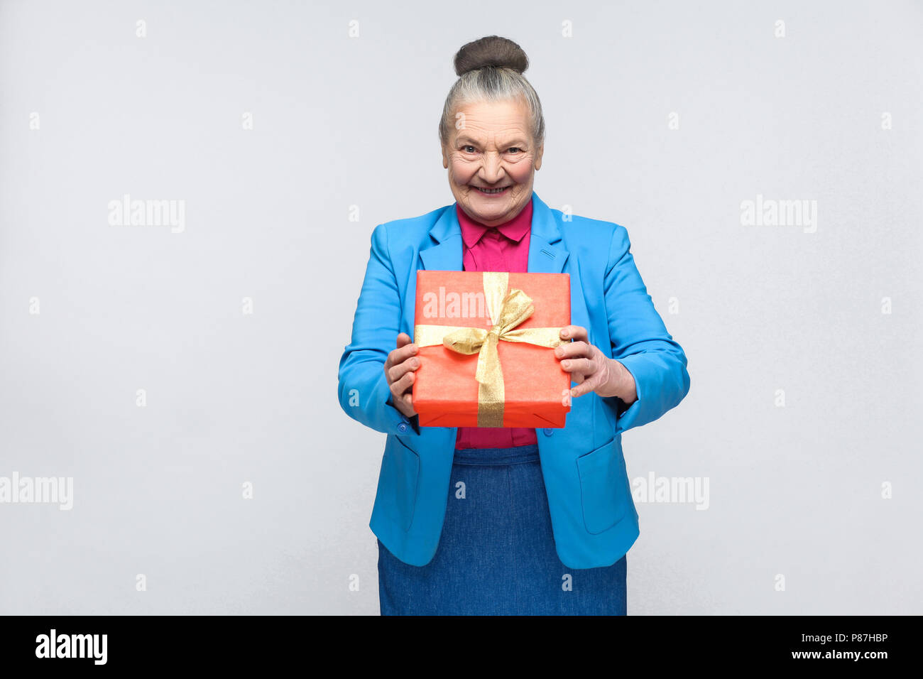 Happiness woman holding red gift box and toothy smiling. Portrait of handsome expressive grandmother in light blue suit with collected gray hair bun h Stock Photo