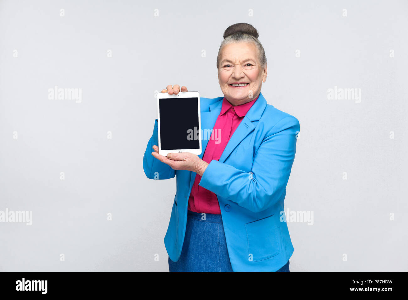 Aged woman holding and showing tablet screen with toothy smiling and satisfied face. Grandmother in light blue suit with collected gray hair bun hairs Stock Photo