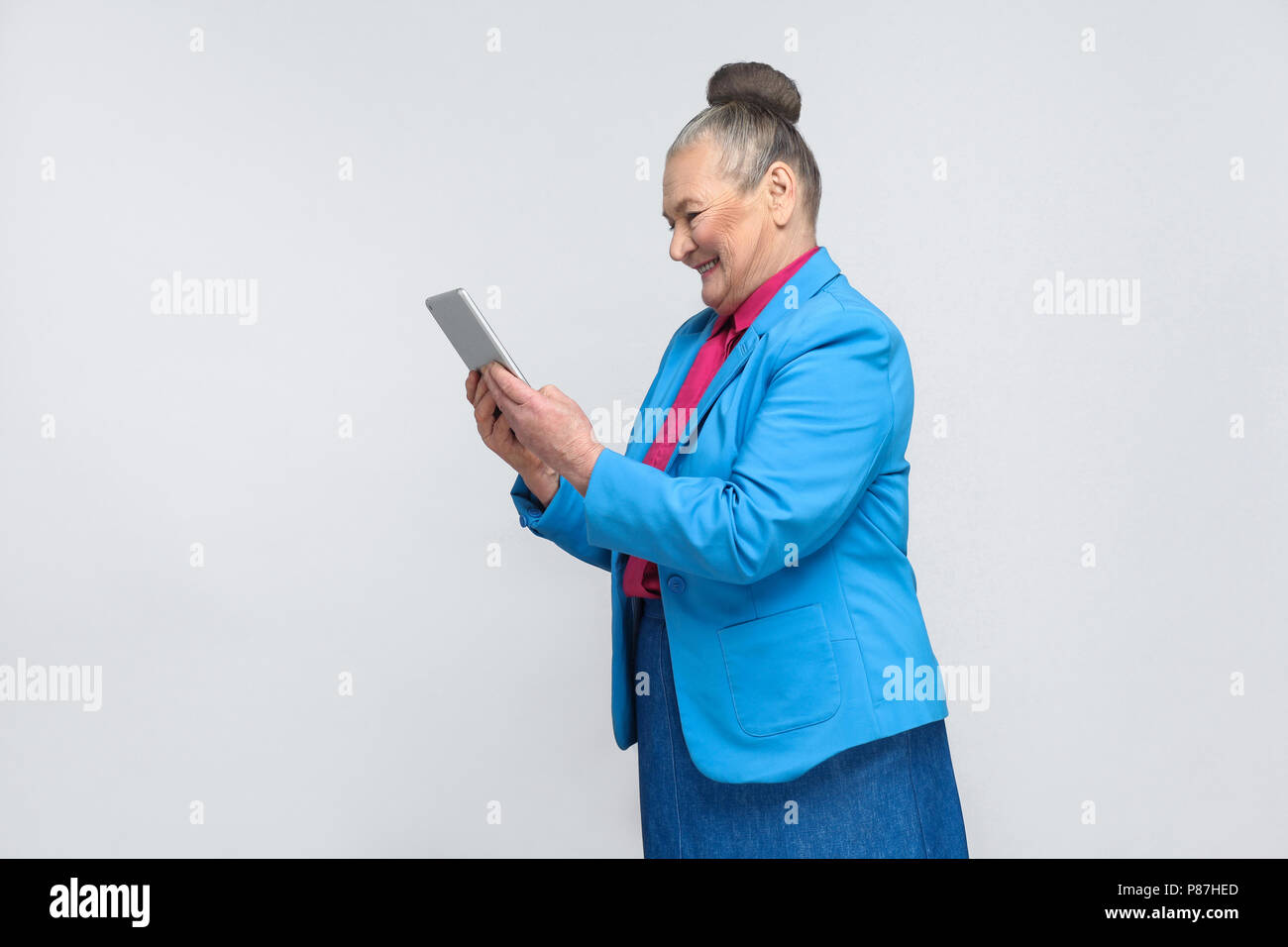 Profile view of aged woman holding tablet and looking at screen with satisfied face and toothy smiling. Grandmother in blue suit with gray hair bun ha Stock Photo
