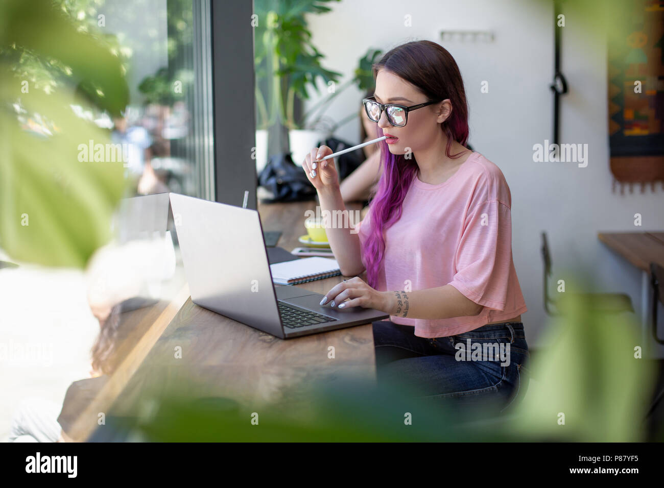 Side view of young businesswoman sitting at table in coffee shop. On table cup of coffee and laptop. In background white wall and window. Stock Photo