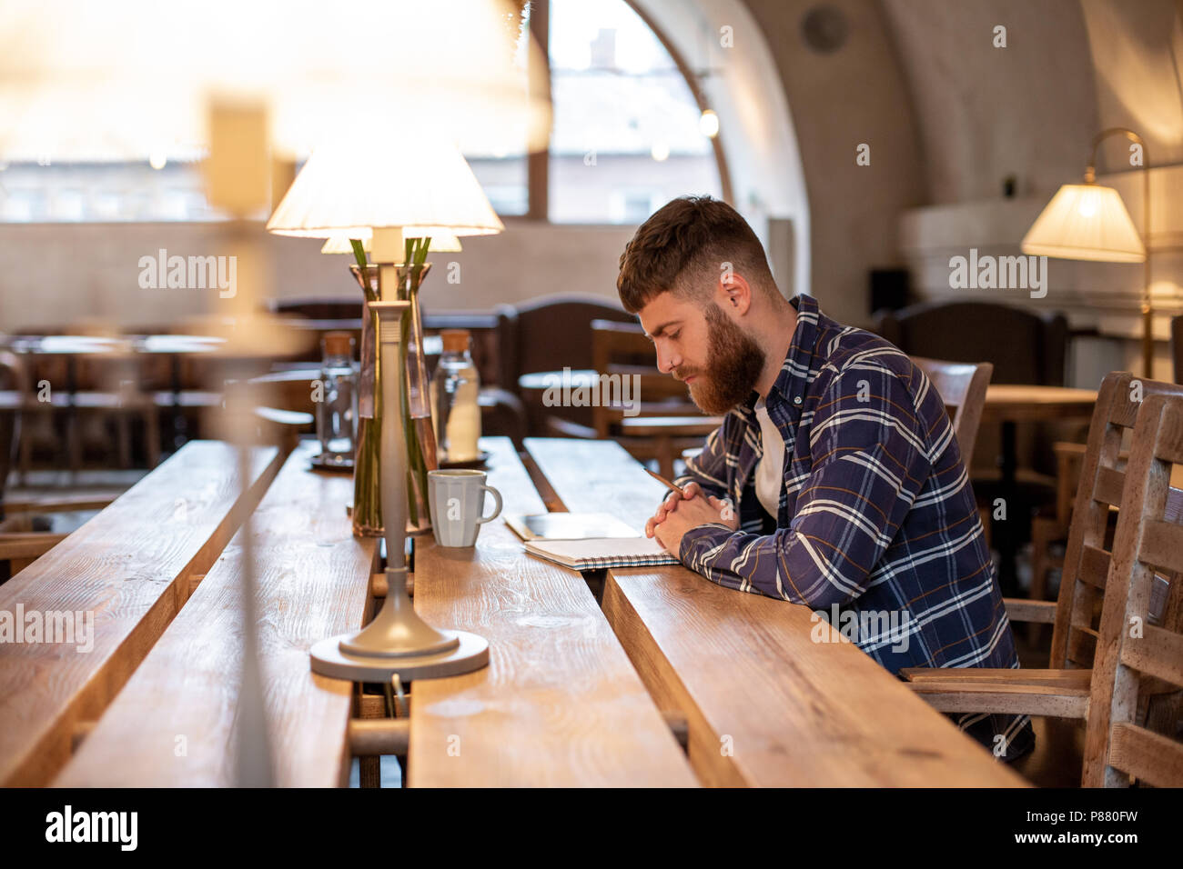Young bearded businessman sits in cafe, home at table and writes in notebook, near lies tablet computer. Man is working, studying. Stock Photo