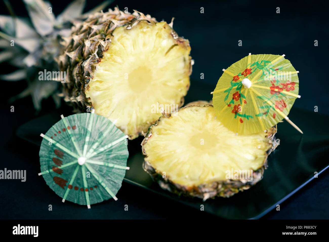 Slices of ripe pineapple on a black plate with decorations for cocktails, umbrella on dark background Stock Photo