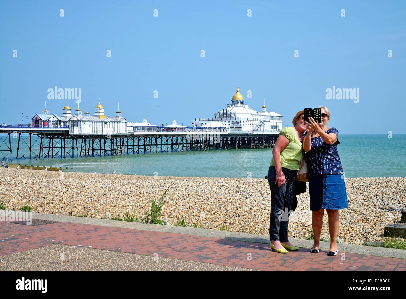 Two elderly women taking a selfie with smart phone on the seafront at Eastbourne with the pier and sea in the background Stock Photo