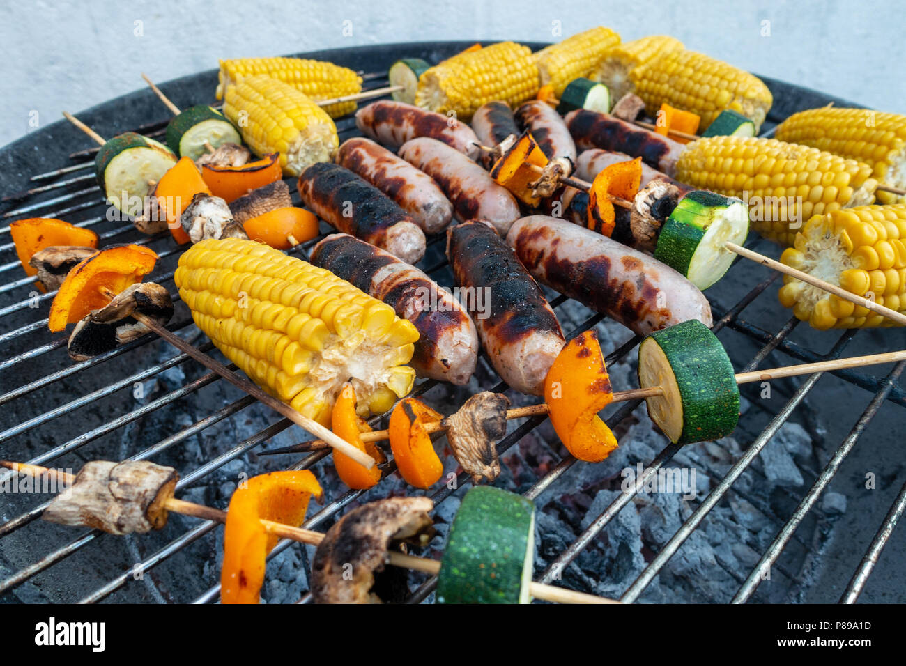 Sausages, corn on the cob and vegetable kebabs of mushroom, pepper and courgette cooking outside on a barbeque. Stock Photo