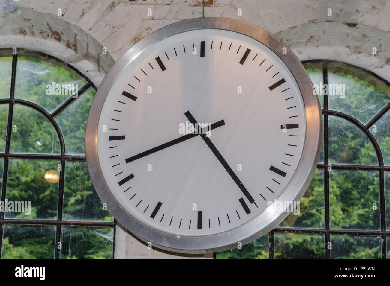 Large clock in front of the window in an industrial factory on a brick wall. Stock Photo