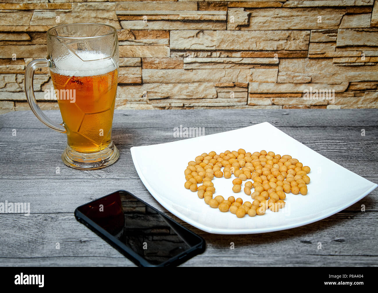On a wooden table is a large glass of beer, a plate of nuts and a phone. Stock Photo