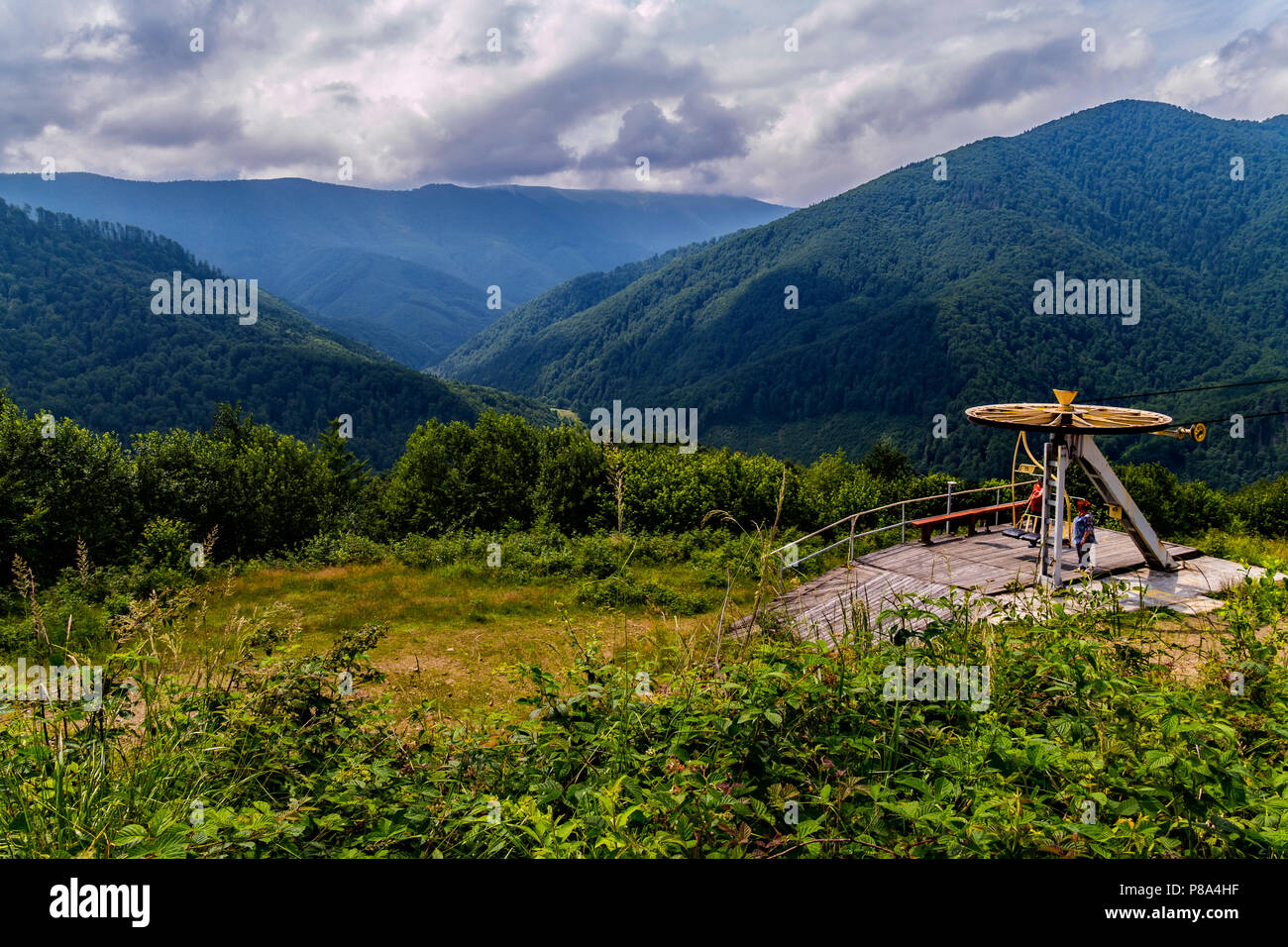 Platform with a lift wheel with people standing on it. With a beautiful view of the mountain slopes covered with green trees going into the distance.  Stock Photo