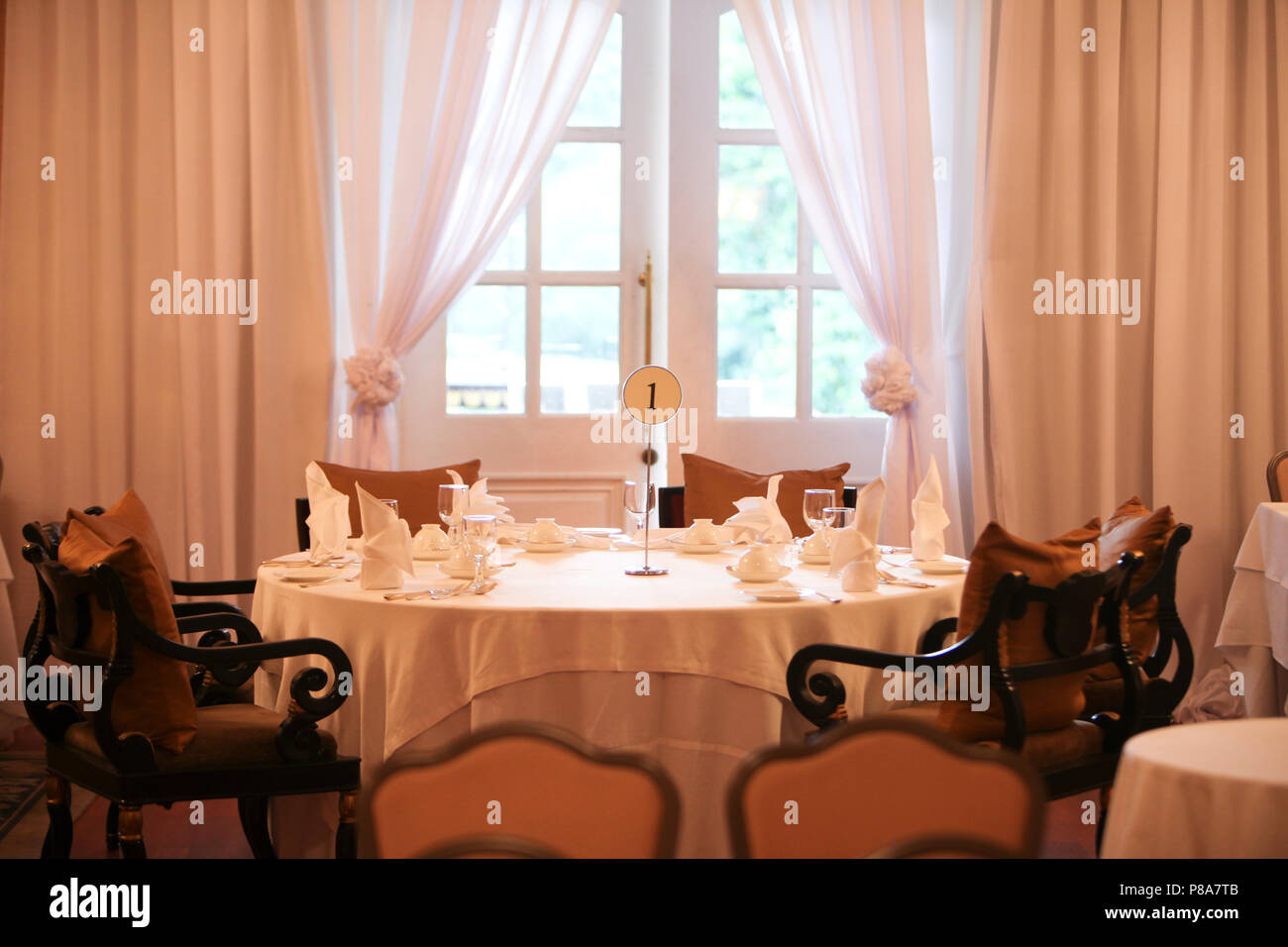 Close-up of a big round numbered table in front of a French window in a festive colonial dining room with white sheer curtains, a white tablecloth and... Stock Photo