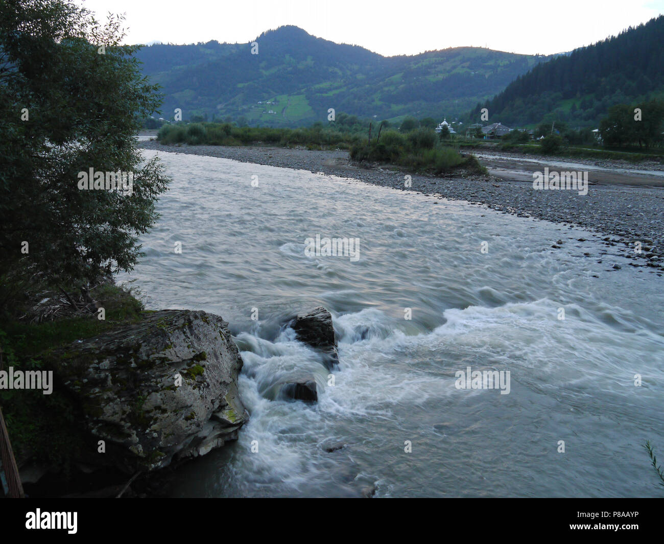 Fast, swift river flow against the backdrop of village houses at the foot of a large mountain . For your design Stock Photo