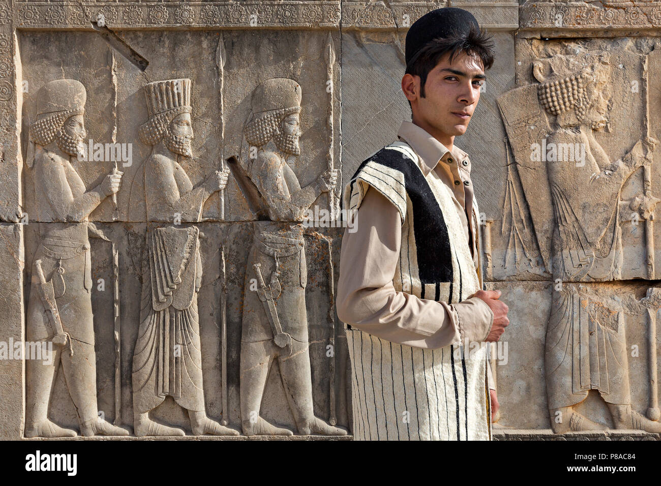 Ethnic Bakhtiari young man in his traditional clothes with Persian reliefs behind him, in Persepolis, Iran. Stock Photo