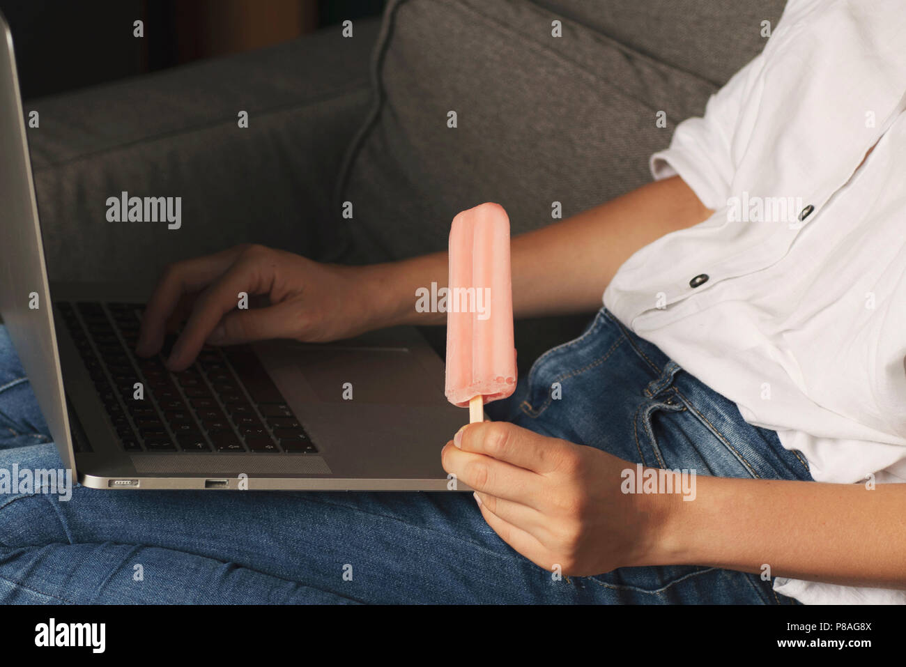 Young woman eating ice cream using laptop computer. Female working on laptop in an outdoor, sitting on sofa at home Stock Photo
