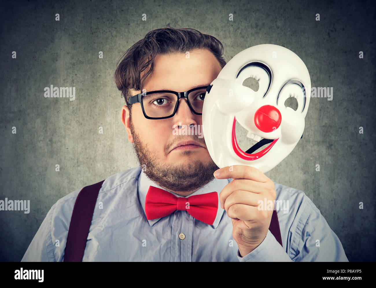 Young bearded man holding happy mask being sad and gloomy while looking at camera on gray background Stock Photo