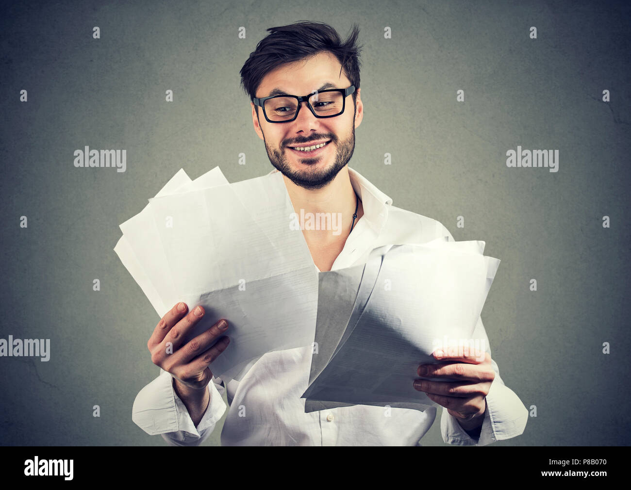 Happy young man in glasses looking through paper documents with good results and smiling on gray background Stock Photo