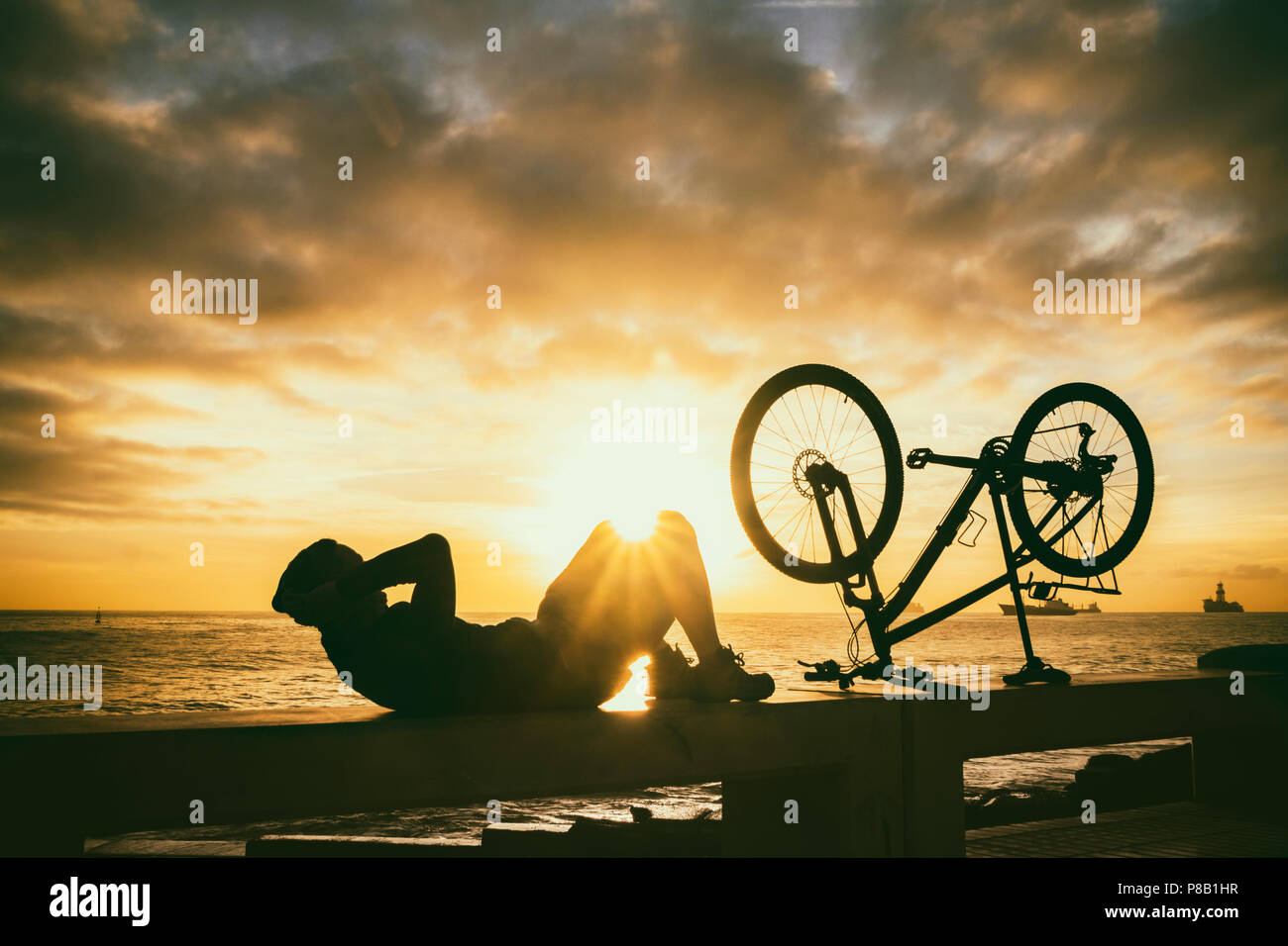 Male Mountain biker doing sit ups on sea wall at sunriise Stock Photo
