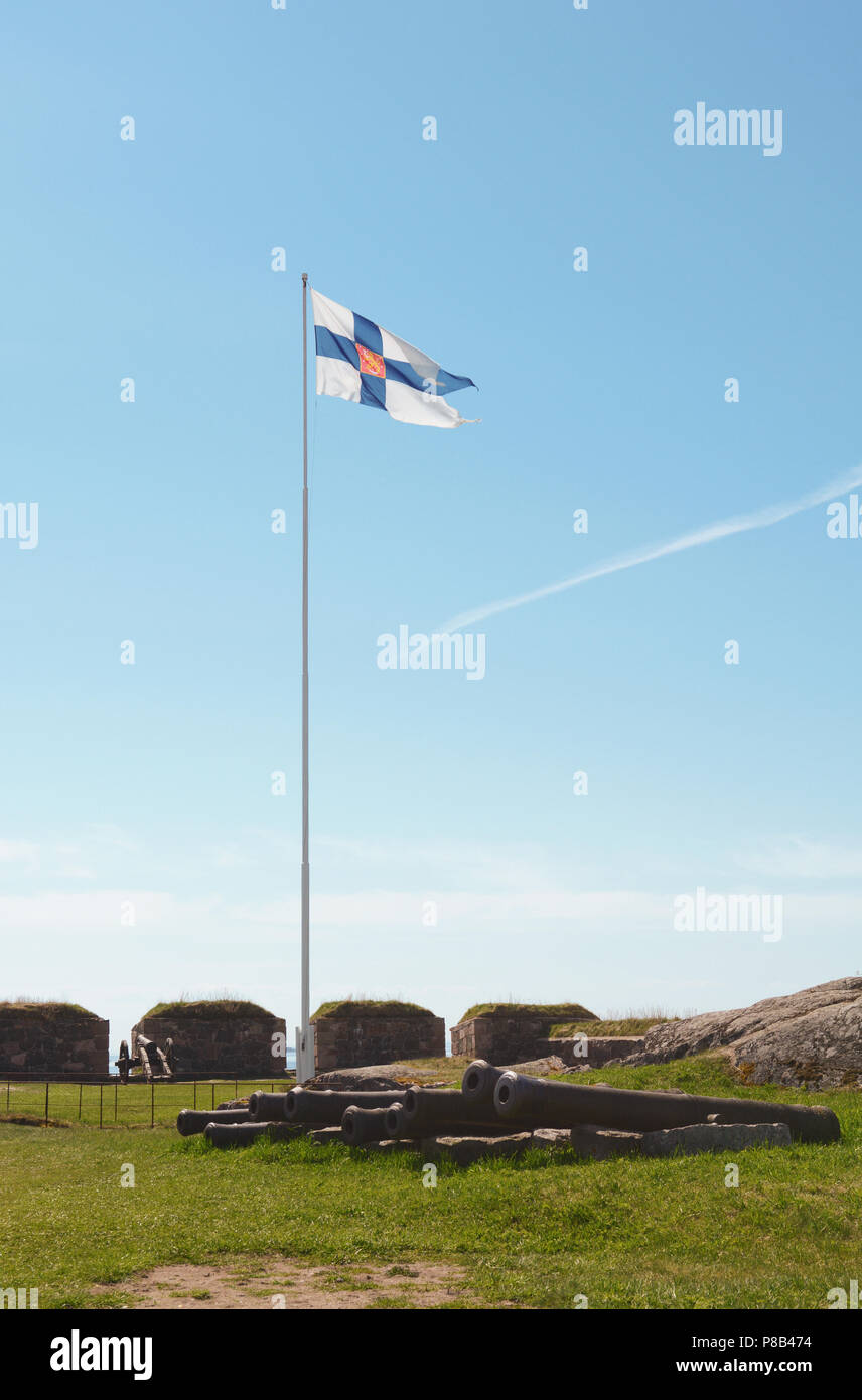 Finland state flag flying high on a flagpole on Suomenlinna sea fortress. A row of disused cannons lies below on the grass. Stock Photo