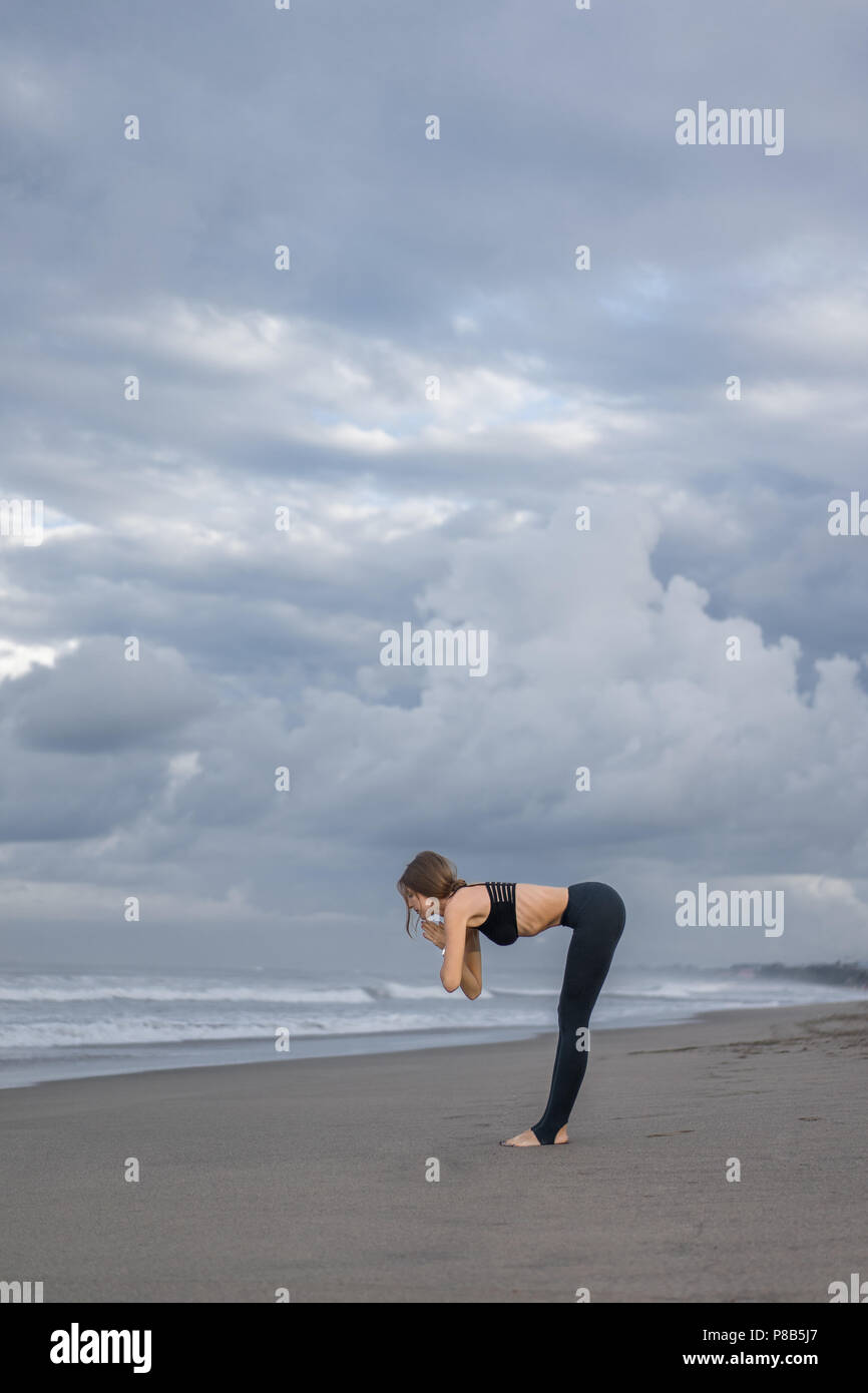 side view of young woman practicing yoga in Halfway Lift pose (Ardha Uttanasana) on seashore Stock Photo