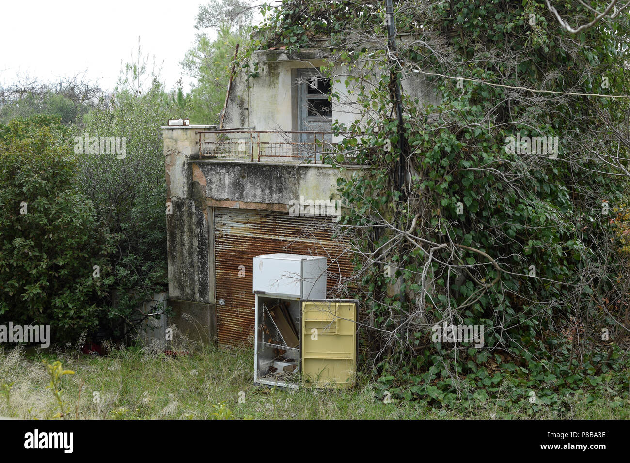 Abandoned house overrun by nature and discarded fridge in garden with overgrown plants. Stock Photo