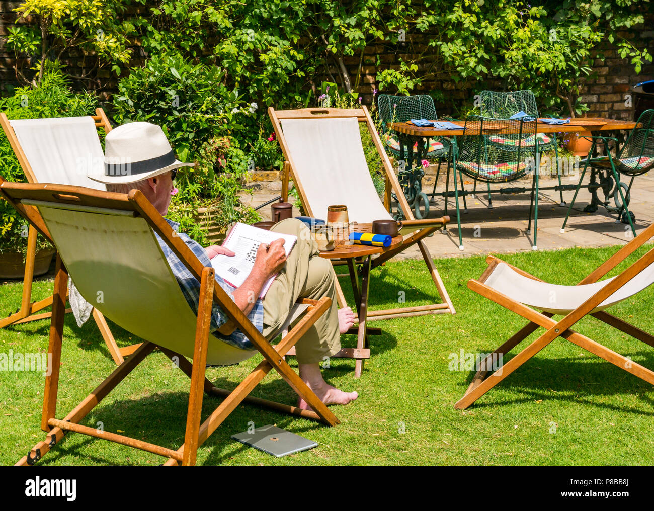 Older man wearing Panama hat solving crossword sitting in deck chair in large garden in Summer heatwave, London, England, UK Stock Photo