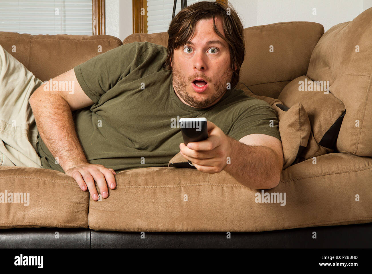 Over weight man watching something shocking on TV. Could it be tabloids, celebrity gossip, a movie, or something else? Stock Photo